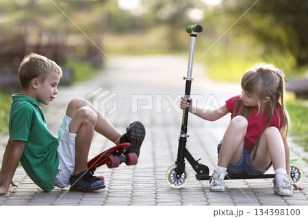 Two cute happy funny laughing young children, brother and sister, having fun on sunny pavement, handsome boy with skateboard and pretty long-haired girl fallen from scooter on blurred background. Two cute happy funny laughing young children, brother and sister, having fun on sunny pavement, handsome boy with skateboard and pretty long-haired girl fallen from scooter on blurred background. 134398100