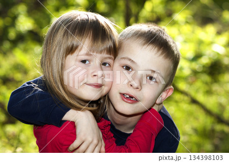 Two cute blond funny happy smiling children siblings, young boy brother embracing sister girl outdoors on bright sunny green bokeh background. Family relation, friendship and love concept. 134398103