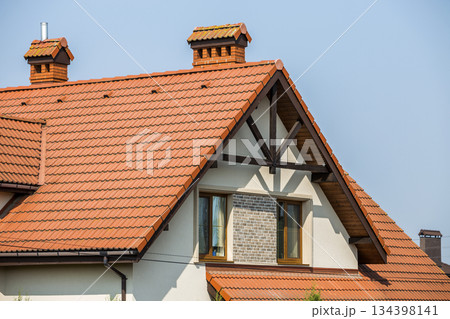 Top of big modern expensive residential house cottage with steep shingled brown roof, high brick chimneys, stucco walls, gutter system and plastic attic windows on blue sky copy space background. Top of big modern expensive residential house cottage with steep shingled brown roof, high brick chimneys, stucco walls, gutter system and plastic attic windows on blue sky copy space background. 134398141
