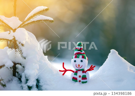 Small funny toy baby snowman in knitted hat and scarf in deep snow outdoors on bright blue and white copy space background. Happy New Year and Merry Christmas greeting card. 134398186