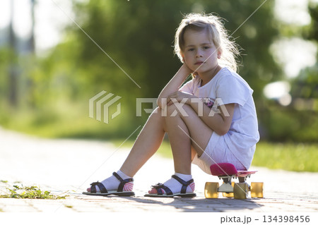 Pretty cute little blond girl in white shorts and T-shirt sitting on pink skateboard and smiling in camera on a light blurred summer background. Children activities and active lifestyle concept. 134398456