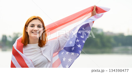 Portrait of happy smiling red haired girl with USA national flag on her shoulders. Positive young woman celebrating United States independence day. Portrait of happy smiling red haired girl with USA national flag on her shoulders. Positive young woman celebrating United States independence day. 134398477
