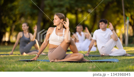 Girl performing Lord of Fishes Pose during group yoga practice outdoors Girl performing Lord of Fishes Pose during group yoga practice outdoors 134398845