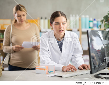 Girl pharmacist works at computer and interprets doctors prescription for pregnant woman client. 134398942