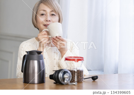 リビングでコーヒー豆を挽いてコーヒーを飲んで寛ぐシニア女性 リビングでコーヒー豆を挽いてコーヒーを飲んで寛ぐシニア女性 134399605