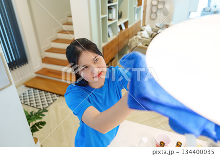 Home Cleaning. Young woman dusting lamp in bright living room. 134400035