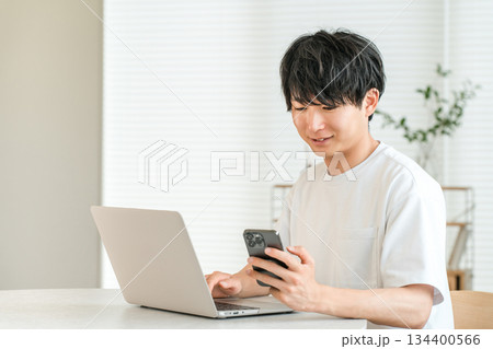 家のリビングでノートパソコンとスマホを使う若いアジア人男性（個人投資家,投資家,投資,資産運用） 134400566