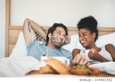 Happy interracial couple, bed and breakfast in relax for morning, bonding or relationship at home. Man and woman smiling with food, coffee or meal relaxing on holiday or weekend together in bedroom Happy interracial couple, bed and breakfast in relax for morning, bonding or relationship at home. Man and woman smiling with food, coffee or meal relaxing on holiday or weekend together in bedroom 134401782