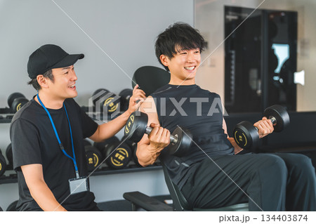 フィットネスジムでダンベルトレーニングを男性とトレーナーの男性スタッフ 134403874
