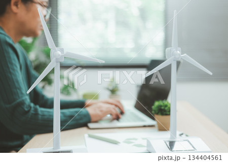 Eco-friendly office scene with person working on laptop near solar panel and wind turbine model, representing green business, renewable energy innovation and sustainable technology workspace. 134404561