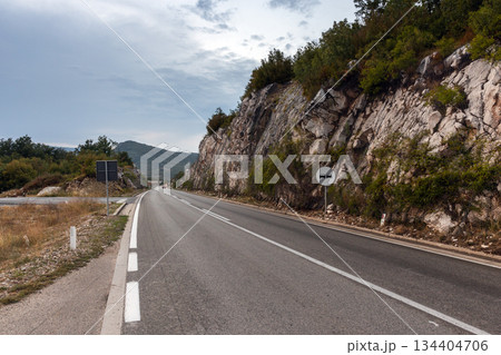 A winding mountain road cuts along a rocky cliffside under a cloudy sky 134404706