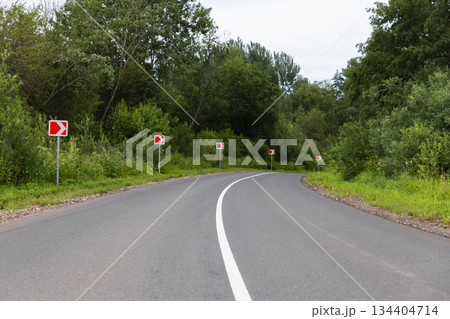 A quiet rural road winds through dense green trees marked by red chevron signs 134404714