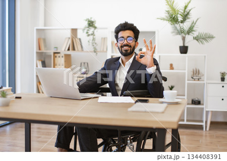 An Indian businessman in a wheelchair smiles while working on his laptop in a modern office setting An Indian businessman in a wheelchair smiles while working on his laptop in a modern office setting 134408391