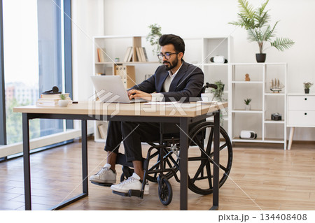 A stylish businessman in a wheelchair works on his laptop in a modern office setting A stylish businessman in a wheelchair works on his laptop in a modern office setting 134408408