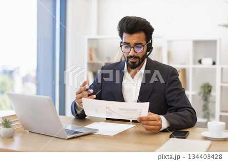 An Indian businessman wearing a headset reviews documents while working on his laptop in a modern office setting An Indian businessman wearing a headset reviews documents while working on his laptop in a modern office setting 134408428