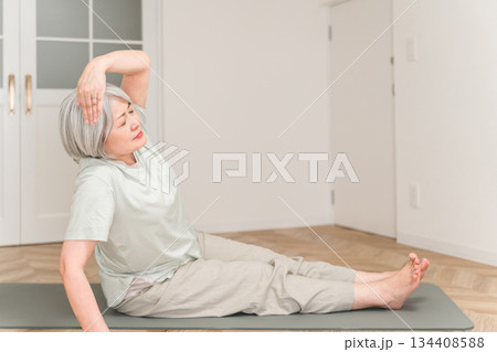 部屋でヨガマットを引いてストレッチ,柔軟体操するグレイヘアーのシニア女性 134408588