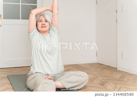 部屋でヨガマットを引いてストレッチ,柔軟体操するグレイヘアーのシニア女性 134408590