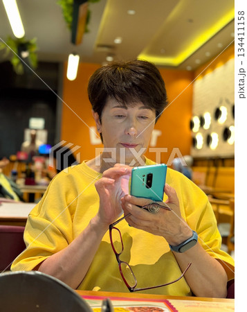 Middle-aged woman using smartphone at restaurant. Casual dining, focused interaction with technology, modern communication. 134411158