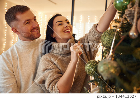 Standing and decorating Christmas tree. Couple is celebrating new year indoors together 134412627