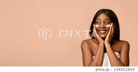 Beautiful african american young lady with face cream or serum on her cheeks. Smiling topless black woman with eye cream on her face, beige studio background, panorama with copy space Beautiful african american young lady with face cream or serum on her cheeks. Smiling topless black woman with eye cream on her face, beige studio background, panorama with copy space 134412819
