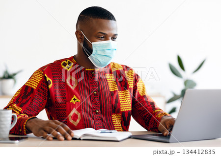 Work During Pandemic. Black Freelancer Guy In Medical Mask Using Laptop At Home, Having Remote Job During Coronavirus Lockdown, African Man In Traditional Clothes Sitting At Desk, Typing On Keyboard Work During Pandemic. Black Freelancer Guy In Medical Mask Using Laptop At Home, Having Remote Job During Coronavirus Lockdown, African Man In Traditional Clothes Sitting At Desk, Typing On Keyboard 134412835