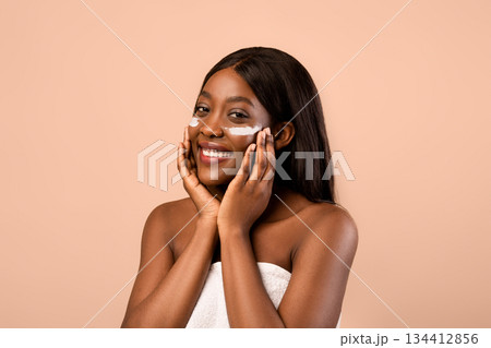 Nude black woman applying white eye cream after shower, beige studio background, closeup. Smiling beautiful african american lady wrapped in towel rubbing under-eyes zone with serum or cream Nude black woman applying white eye cream after shower, beige studio background, closeup. Smiling beautiful african american lady wrapped in towel rubbing under-eyes zone with serum or cream 134412856