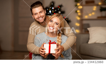 Cheerful young couple hugging and holding gift box, exchanging with Christmas presents at home, empty space. Smiling man and woman presenting xmas gifts to each other, celebrating winter holidays Cheerful young couple hugging and holding gift box, exchanging with Christmas presents at home, empty space. Smiling man and woman presenting xmas gifts to each other, celebrating winter holidays 134412860