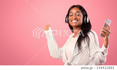 Lovely black lady with headphones and mobile device listening to music and dancing on pink studio background. Beautiful African American woman moving to her favorite song with closed eyes 134412883