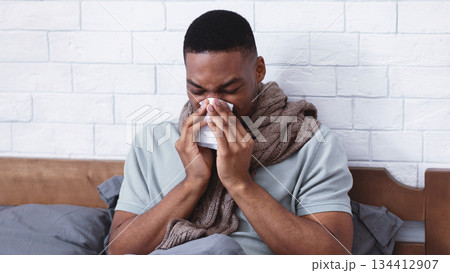 Sick African Man Blowing Runny Nose In Paper Tissue Suffering From Rhinitis Disease Sitting In Bed At Home. Ill Black Guy Sneezing Having Sinusitis And Cold Symptoms Concept. Free Space Sick African Man Blowing Runny Nose In Paper Tissue Suffering From Rhinitis Disease Sitting In Bed At Home. Ill Black Guy Sneezing Having Sinusitis And Cold Symptoms Concept. Free Space 134412907