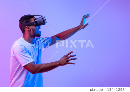 Modern Gaming. Amazed Black Man Touching Air While Experiencing Virtual Reality In VR Glasses, Excited Young Black Guy Enjoying Playing Video Games, Standing Under Neon Light Over Purple Background Modern Gaming. Amazed Black Man Touching Air While Experiencing Virtual Reality In VR Glasses, Excited Young Black Guy Enjoying Playing Video Games, Standing Under Neon Light Over Purple Background 134412984