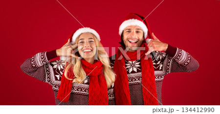 Joyful young couple in Christmas sweaters and Santa hats showing CALL ME gesture on red background 134412990