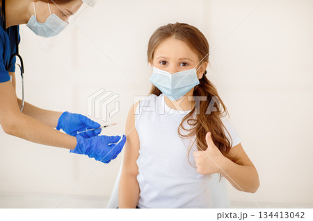 Little girl in face mask gesturing thumb up while receiving covid-19 vaccine injection at health centre. Cute child approving of coronavirus immunization, being protected against global virus 134413042