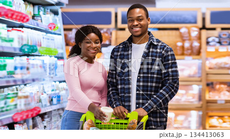 Portrait of positive black couple with trolley full of products looking at camera and smiling at huge supermarket. Happy African American woman and man buying groceries at mall Portrait of positive black couple with trolley full of products looking at camera and smiling at huge supermarket. Happy African American woman and man buying groceries at mall 134413063