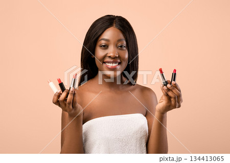 Closeup of smiling beautiful young black woman wrapped in towel holding two lipsticks in her hands, posing over beige background. Professional cosmetics makeup, lipstick, lip-gloss concept Closeup of smiling beautiful young black woman wrapped in towel holding two lipsticks in her hands, posing over beige background. Professional cosmetics makeup, lipstick, lip-gloss concept 134413065