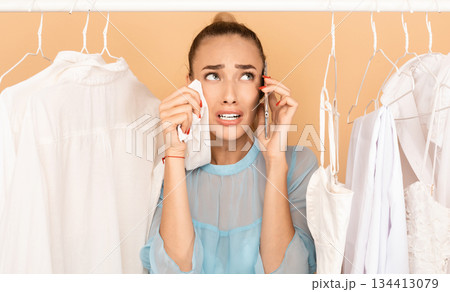 I Have Nothing To Wear. Portrait of upset stressed woman talking on mobile phone and crying, wiping tears with shirt, having panic, standing in rack of clothes between hangers I Have Nothing To Wear. Portrait of upset stressed woman talking on mobile phone and crying, wiping tears with shirt, having panic, standing in rack of clothes between hangers 134413079