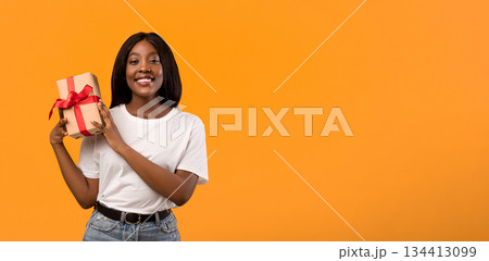Happy african american young woman holding gift box next to her ear, wondering what is inside, orange studio background, panorama with copy space. Birthday gift, present, surprise concept 134413099
