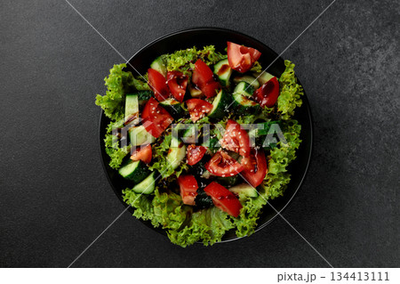 Vegetable salad of tomatoes, cucumbers, onions, lettuce dressed with aromatic dressing in a black plate on a gray background 134413111