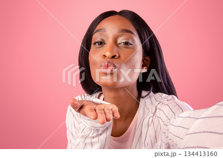 Attractive young black woman taking selfie, blowing air kiss on pink studio background. Lovely African American lady posing to mobile camera and making photo of herself Attractive young black woman taking selfie, blowing air kiss on pink studio background. Lovely African American lady posing to mobile camera and making photo of herself 134413160