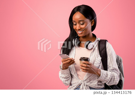 Young black lady using cellphone and holding takeout coffee on pink studio background. African American student with backpack and headphones browsing web on mobile device 134413176