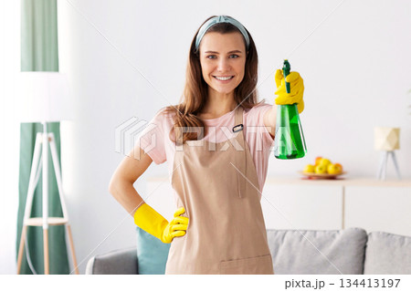 Full length portrait of young maid in apron and rubber gloves pointing detergent spray at camera and smiling in clean living room, copy space. Professional cleaning service concept Full length portrait of young maid in apron and rubber gloves pointing detergent spray at camera and smiling in clean living room, copy space. Professional cleaning service concept 134413197
