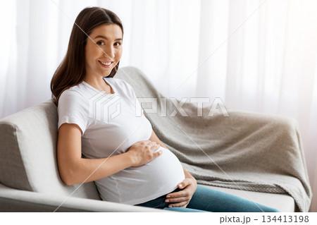 Portrait of beautiful young pregnant lady in casual clothes sitting on couch and tenderly embracing belly, happy millennial expectant woman relaxing in living room at home and smiling at camera 134413198