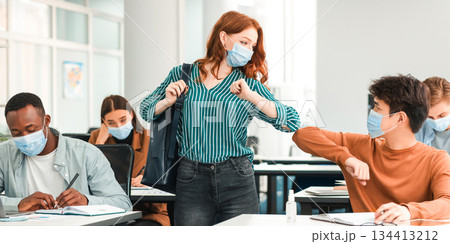 Stop Spreading Virus Concept. Smiling diverse classmates wearing protective face masks greeting each other and bumping elbows at classroom. Woman standing with backpack, guy sitting at table Stop Spreading Virus Concept. Smiling diverse classmates wearing protective face masks greeting each other and bumping elbows at classroom. Woman standing with backpack, guy sitting at table 134413212