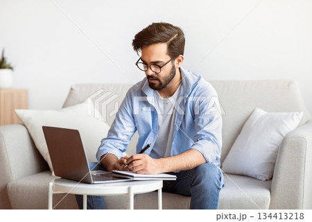 Online Education. Young Eastern Man Study With Laptop At Home And Taking Notes, Millennial Arab Guy In Eyeglasses Watching Online Webinar, Sitting On Couch With Computer, Enjoying Distance Learning Online Education. Young Eastern Man Study With Laptop At Home And Taking Notes, Millennial Arab Guy In Eyeglasses Watching Online Webinar, Sitting On Couch With Computer, Enjoying Distance Learning 134413218