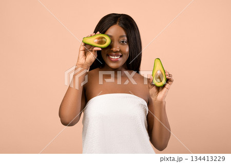 Happy beautiful black woman with long hair wrapped in towel covering eyes with half of avocado over beige studio background. Youth, beauty, anti-aging organic recipes, healthy diet concept 134413229