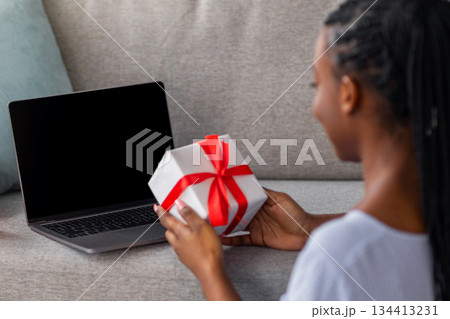 Back view of black lady with dreadlocks holding present, using laptop with empty screen, african american young woman having Birthday party online, sitting on floor in front of notebook 134413231