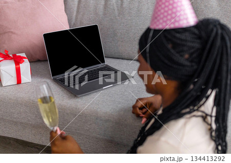 Remote birthday celebration during self-isolation. Young african american woman with glass of champagne and gift box usign laptop with blank screen, wearing party hat, home interior 134413292