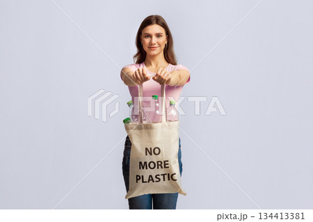 No More Plastic. Happy Young Lady Carrying Eco Tote Bag With Empty Bottles For Recycling, Smiling Nature-Lover Woman Promoting Zero-Waste Living, Posing Over Light Studio Background, Copy Space 134413381