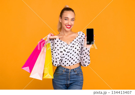 Ordering Online. Portrait of smiling woman holding colorful shopping bags and showing cell phone with blank black screen to camera, standing isolated over pastel orange studio wall background 134413407