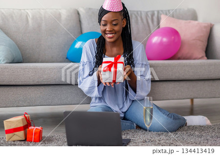 Happy young black woman with party hat holding present and smiling at laptop screen, sitting on floor by couch in living room at home, having online Birthday party during COVID-19 pandemic, copy space Happy young black woman with party hat holding present and smiling at laptop screen, sitting on floor by couch in living room at home, having online Birthday party during COVID-19 pandemic, copy space 134413419
