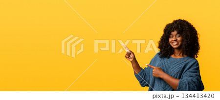Happy black young woman in knitted green sweater pointing at copy space and smiling, yellow studio background, panorama. Cute african american lady showing empty space for advertising 134413420